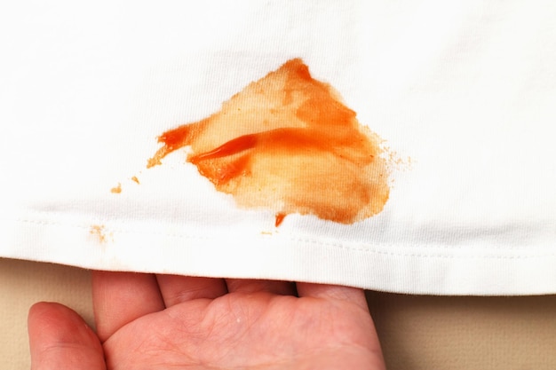 A mão da mulher segura roupas de camisa branca com mancha de ketchup em um fundo bege