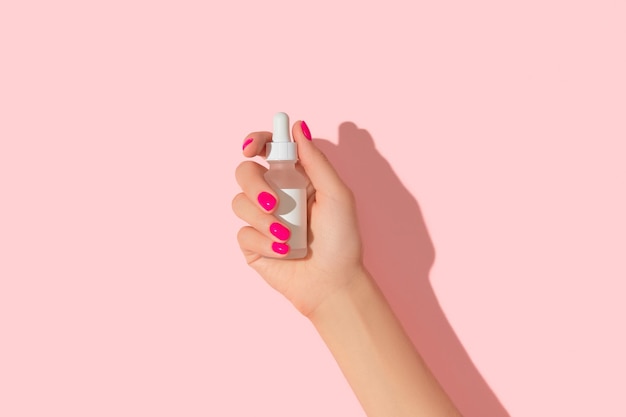 Foto a mão da mulher segurando o soro conta-gotas no conceito de tratamento de beleza de autocuidado de fundo rosa pastel