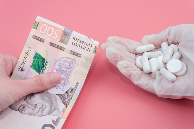 A mão dá uma conta de quinhentos hryvnia para uma mão em uma luva médica com um comprimido em um fundo rosa. Aumento do preço dos medicamentos.