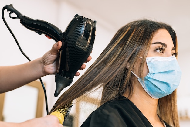 A mão de um cabeleireiro carregando um secador de cabelo secando o cabelo de uma linda garota caucasiana com uma máscara facial por causa do coronavírus. A menina tem cabelo comprido