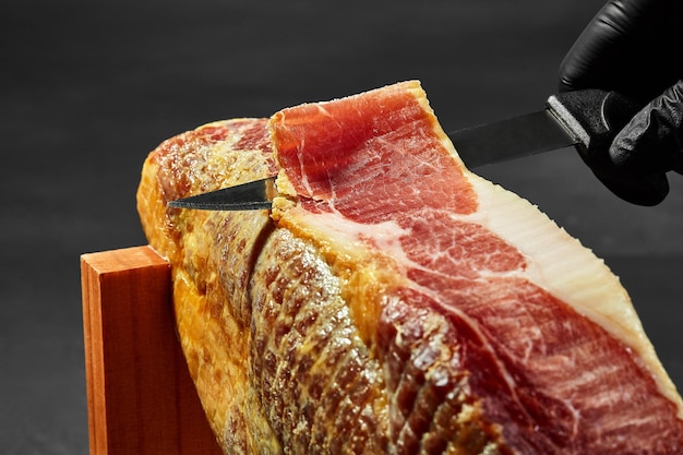 A mão de um cozinheiro em uma luva preta está cortando um pedaço de jamon com uma faca de uma perna de porco em uma barraca de jamonera Tiro aproximado Vista lateral