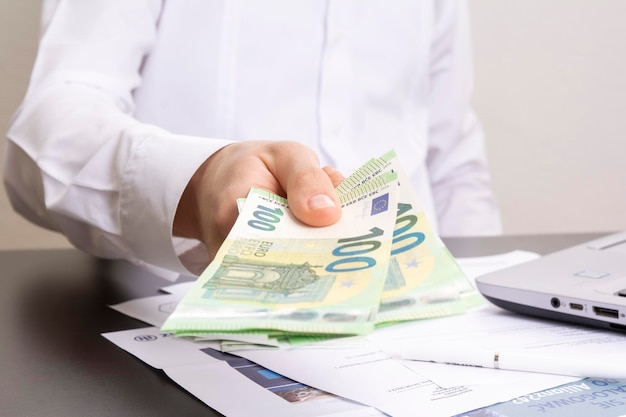 A mão de um homem segura notas de cem euros contra o fundo de uma mesa de escritório com um computador