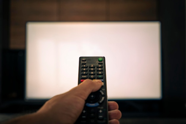 A mão de um homem segura o controle remoto na frente de uma grande tela de TV branca desfocada controle remoto para televisão