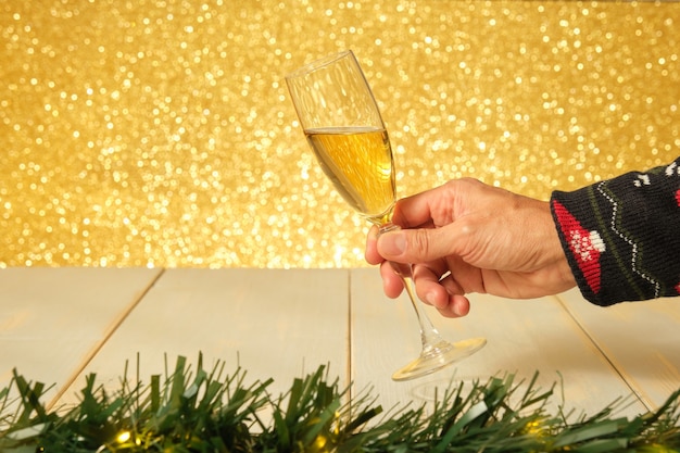 A mão de um homem segurando uma taça de champanhe em uma mesa de madeira branca e envelhecida com um fundo dourado brilhante