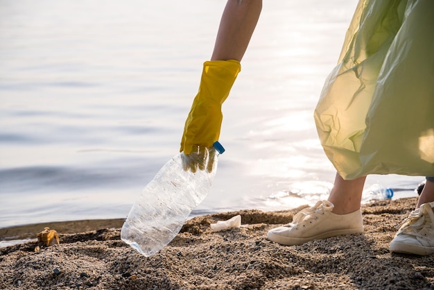 Foto a mão de um voluntário em uma luva de borracha amarela remove garrafas plásticas da margem arenosa da lagoa. recolha e tratamento de resíduos. subbotnik organizado. fechar-se.