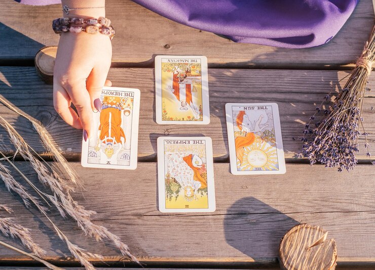 A mão de uma mulher com unhas roxas coloca uma das quatro cartas de tarô espalhadas em uma superfície de madeira ao lado de espigas e alfazema