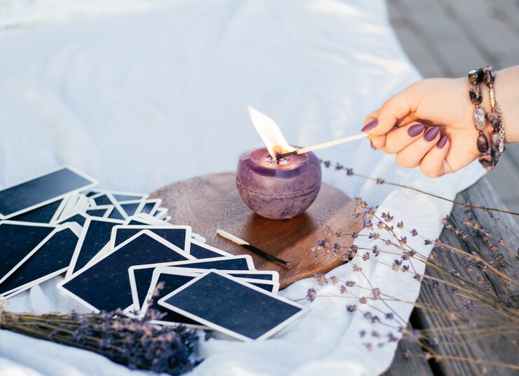 A mão de uma mulher em uma pulseira com unhas compridas roxas acende uma vela com um fósforo, ao lado do baralho de cartas e flores de lavanda
