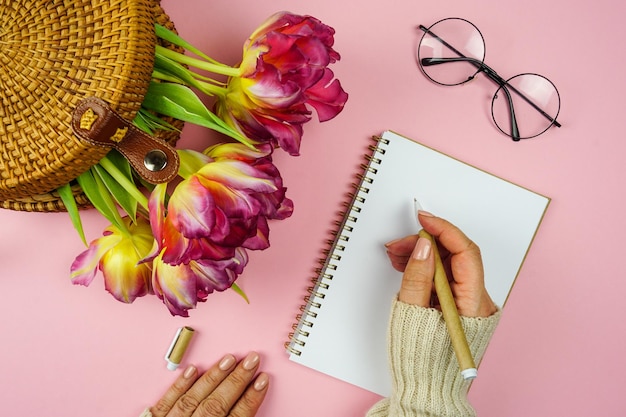 A mão de uma mulher escreve planos em um caderno, há tulipas em um saco de vime na mesa