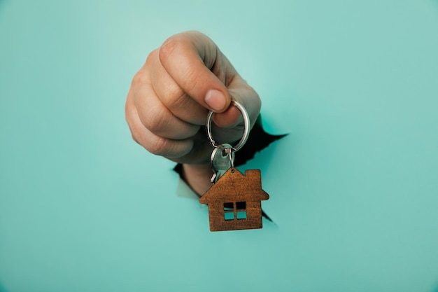 A mão do homem com a chave da casa através de um buraco no fundo azul Conceito de venda e aluguel de casa