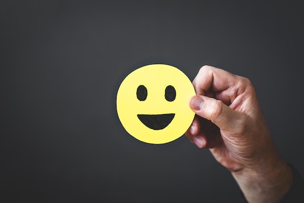 A mão do homem em um terno de negócios segura uma carinha amarela com um ícone de rosto feliz em um fundo escuro O conceito de satisfação do cliente ou feedback positivo