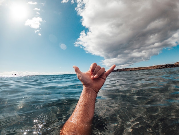 A mão do homem no sinal de surfe na água azul do oceano