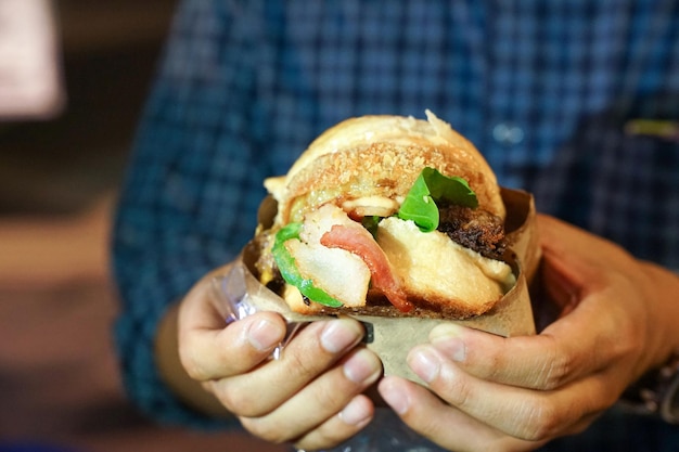 A mão do homem segura um grande bacon de hambúrguer e está pronto para mordê-lo na boca no evento de foodtruck da Tailândia