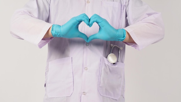A mão do médico faz sinal cardíaco de amor com um estetoscópio. A mão usa a luva médica azul e um vestido de manga comprida em fundo branco.