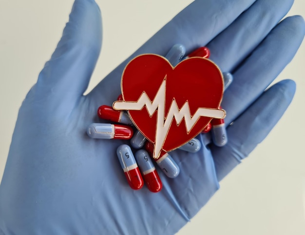 A mão do médico na luva segura o símbolo do coração vermelho e as pílulas para o coração