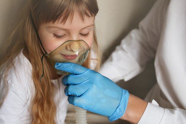 A mão do médico nas luvas segura um inalador de vapor de máscara de oxigênio para uma menina, tratamento para asma, respirando através de um nebulizador de vapor.