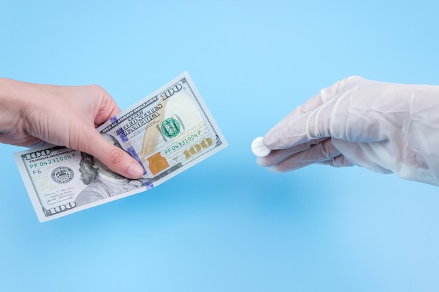 A mão estende uma nota de cem dólares para a mão em uma luva médica com um comprimido em um fundo azul. Aumento do preço dos medicamentos