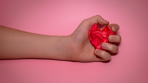 A mão segura o coração de papel amassado vermelho no fundo rosa