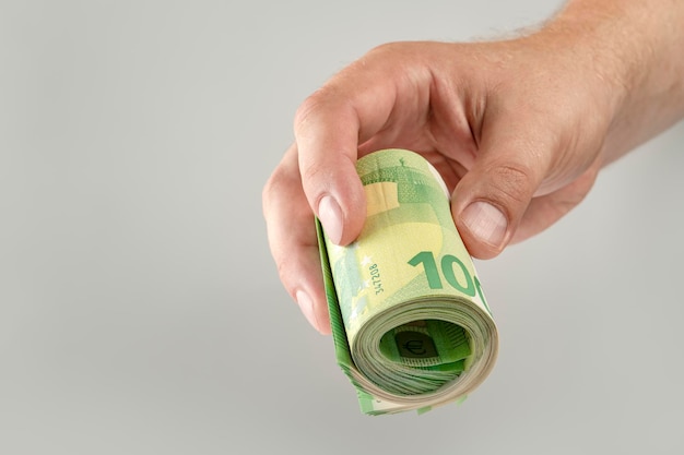 A mão segura um rolo de notas de 100 euros Notas de euro enroladas na mão de um homem branco sobre um fundo cinza O conceito de assistência financeira empréstimo de compra de imóveis ou pagamento de seguro