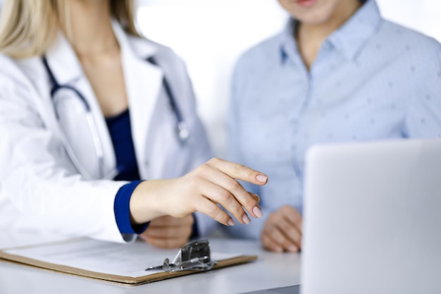 A médica desconhecida e seu paciente estão discutindo o exame de sangue do paciente, enquanto estão sentados juntos na mesa do gabinete de uma clínica. Médica, com estetoscópio, está usando um computador portátil