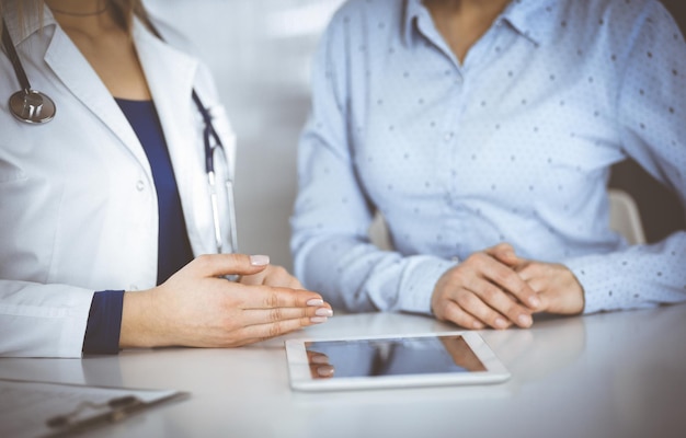 A médica desconhecida está mostrando a sua paciente uma descrição de medicação, enquanto está sentada junto à mesa do gabinete de uma clínica. Médica está usando um tablet de computador e um estetoscópio