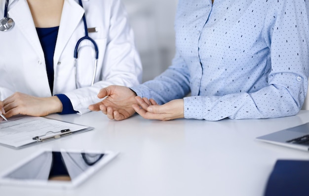 A médica desconhecida está ouvindo seu paciente, enquanto está sentado na mesa do gabinete de uma clínica. Médica com estetoscópio está escrevendo na área de transferência, close-up. Médico perfeito