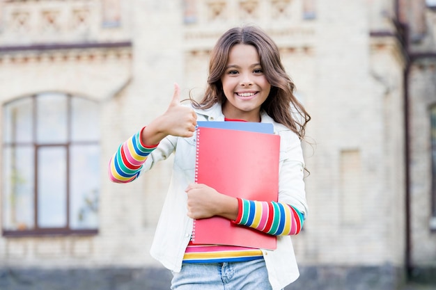 A melhor lição de sempre Criança feliz dando os polegares para a aula de literatura Menina sorrindo com livros de aula ao ar livre A lição pode ser divertida