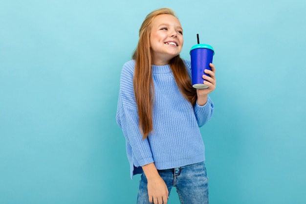 A menina adolescente caucasiano bonita com cabelo castanho no hoody azul bebe o café com o copo azul isolado no azul