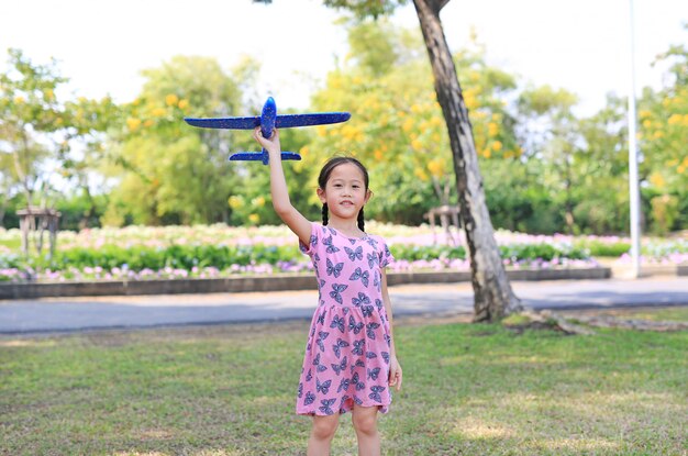 A menina alegre levanta um avião azul do brinquedo que voa no ar no jardim ao ar livre.