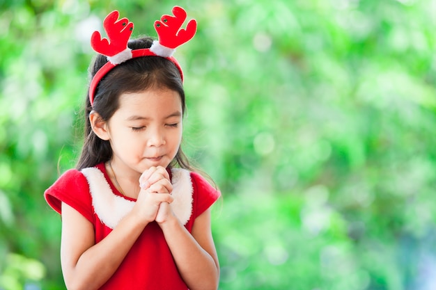 A menina asiática bonito no vestido do natal fechou seus olhos e dobrou sua mão na oração