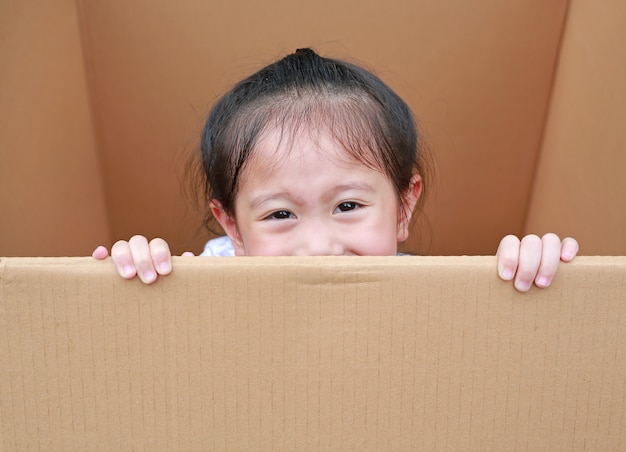 A menina asiática pequena da criança que joga o peekaboo e encontra-se na caixa de cartão grande.