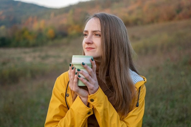 A menina bonita bebe o chá ou o café do copo no fundo da natureza do outono A mulher feliz relaxa fora