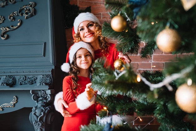A menina bonita com a mãe está decorando uma árvore de Natal na casa. Família feliz.