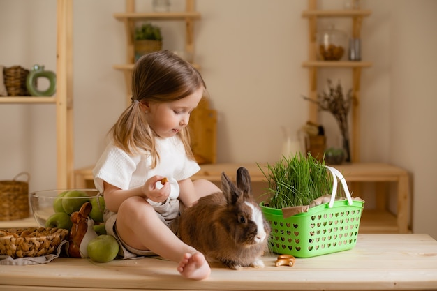 A menina bonitinha na cozinha de madeira da casa alimenta o coelho com grama fresca