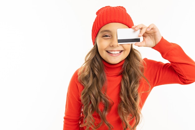 A menina caucasiano feliz no chapéu vermelho e hoody prende um cartão de crédito isolado no branco