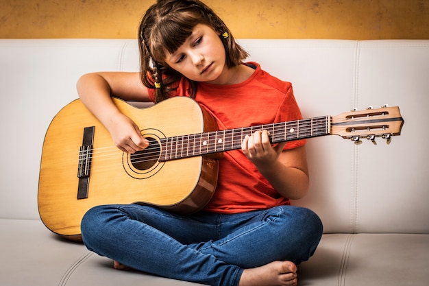 A menina concentrada está praticando com violão senta-se no sofá em casa