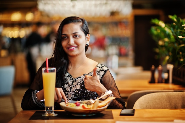 A menina consideravelmente indiana no vestido preto do saree levantou no restaurante, sentando-se na tabela com suco e salada. Mostre o polegar para cima.