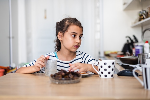 A menina criança feliz come fruta tâmara na cozinha da casa de verão