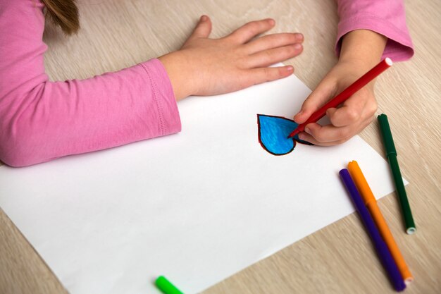 A menina da criança entrega o desenho com coração azul dos pastéis coloridos dos lápis no Livro Branco. Arte educação, conceito de criatividade.