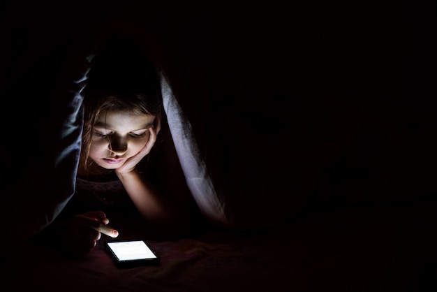 Foto a menina de 9 anos à noite está coberta com um cobertor e está olhando para um smartphone.