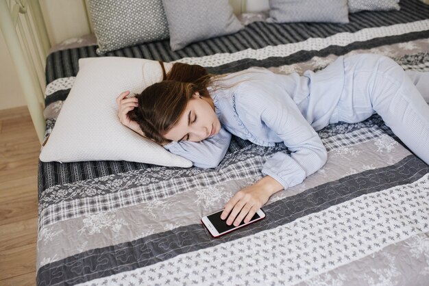 A menina de pijama na cama no quarto dela. Interior cinza-branco elegante. Ela adormeceu com o telefone na mão. Aguardando uma ligação.