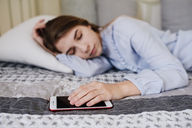 A menina de pijama na cama no quarto dela. Interior cinza-branco elegante. Ela adormeceu com o telefone na mão. Aguardando uma ligação.