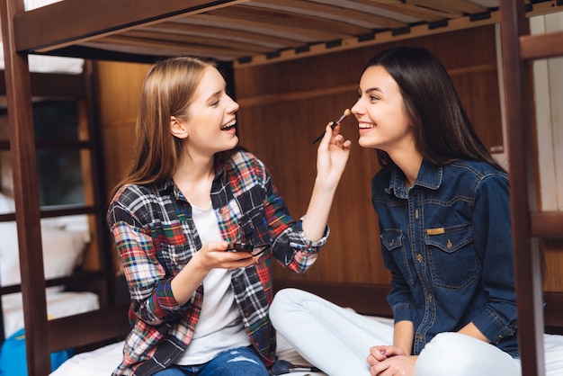 A menina de sorriso senta-se na cama e pinta-se a amiga.