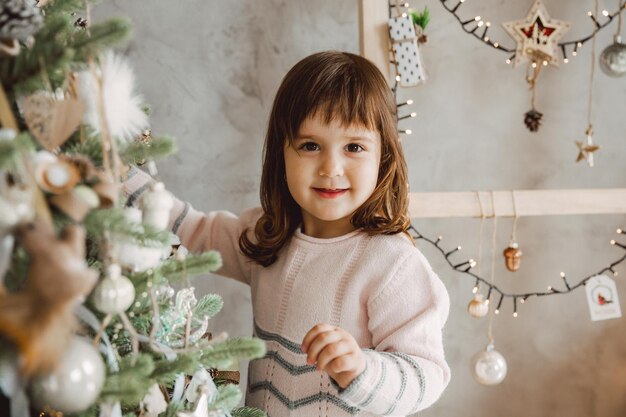 A menina decora a árvore de natal com brinquedos. um lindo bebê está se preparando em casa para a celebração do natal.