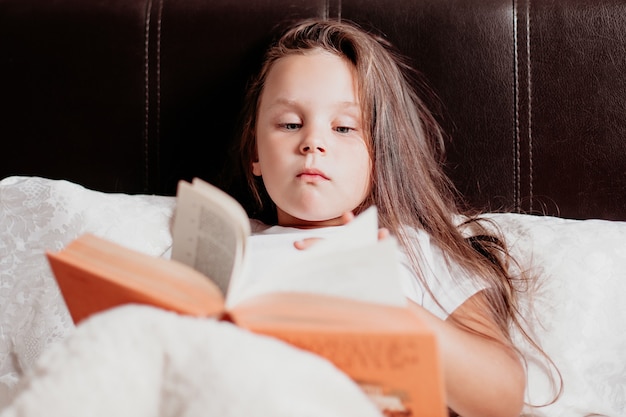 A menina deita-se em uma cama branca e lê um livro laranja, conforto do lar e autoeducação.