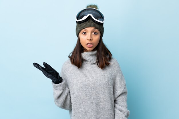 A menina do esquiador da raça misturada com vidros do snowboard sobre a parede azul isolada que faz dúvidas gesticula