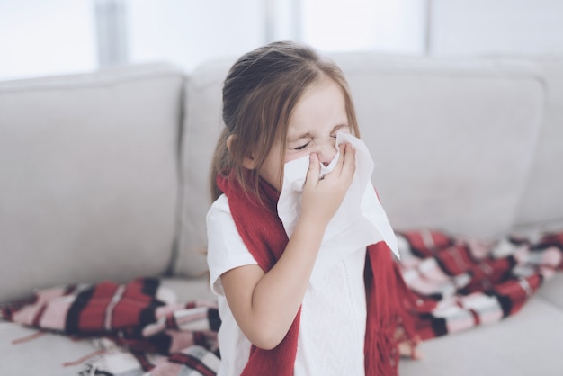 A menina doente senta-se em um sofá branco envolvido em um lenço vermelho.