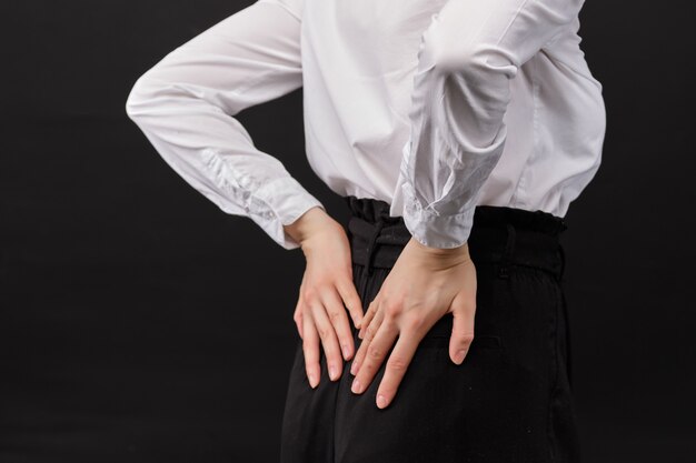 Foto a menina em uma camisa branca massageia a parte inferior das costas em um fundo preto.