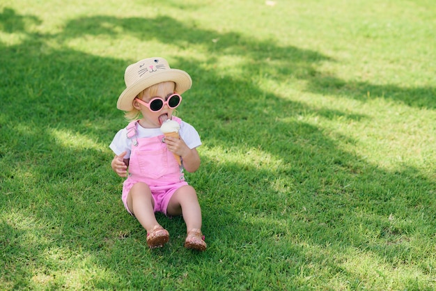 A menina engraçada da criança feliz que veste macacões cor-de-rosa do verão, chapéu e óculos de sol cor-de-rosa senta-se em um gramado verde come o gelado branco de baunilha em um jardim ensolarado.