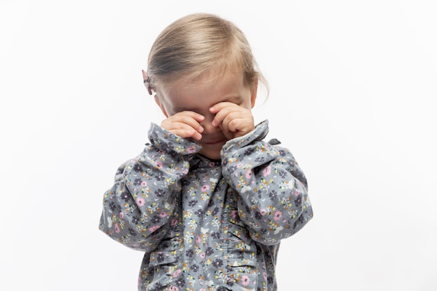 A menina esfrega os olhos com as mãos. Criança em um vestido com estampa floral. Fechar-se. Fundo branco.