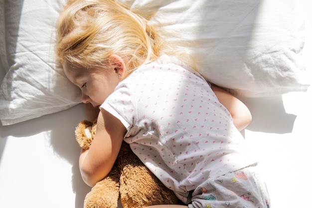 A menina está abraçando um urso de brinquedo macio em uma cama branca Vista superior plana